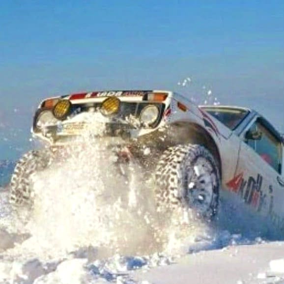 Close-up of Wissam Akoury's Niva driving through deep snow, with snow spraying around its front wheels and additional rally lights mounted above the windscreen.