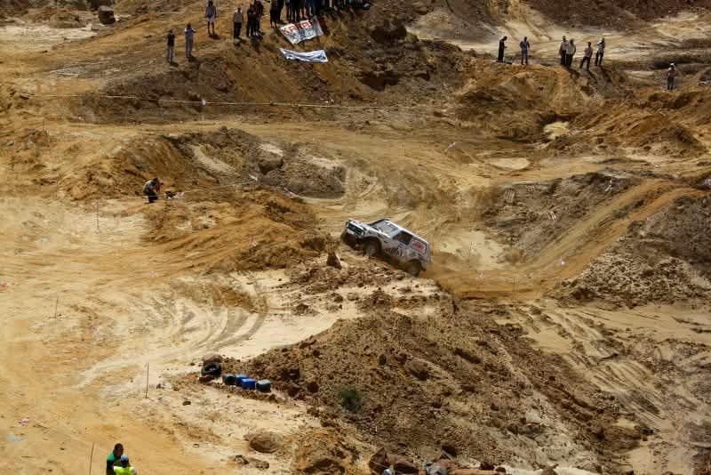 A wide view of the off-road course in a sandy quarry, showing Wissam Akoury's Niva making its way across a deeply rutted slope while spectators watch from higher ground.