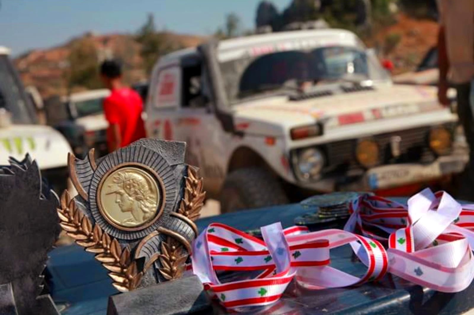A trophy and several medals with Lebanese flag-coloured ribbons displayed on a car hood, with Wissam Akoury's competition Niva visible in the background.