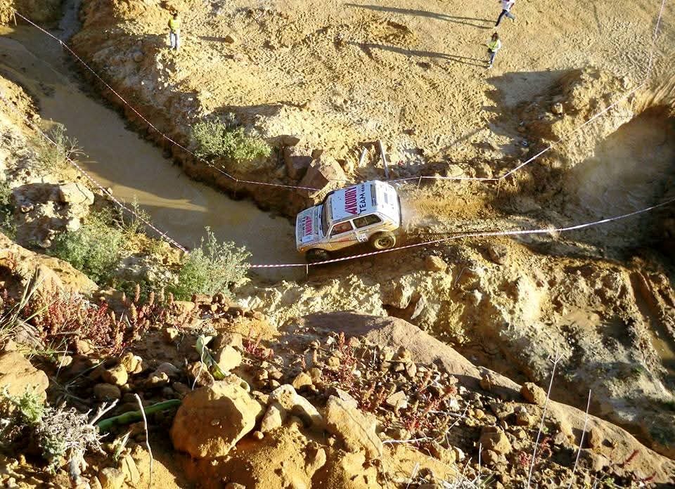 An overhead view of Wissam Akoury's Niva crossing a water-filled trench between rocky banks. Course markers and marshals are visible near the crossing.